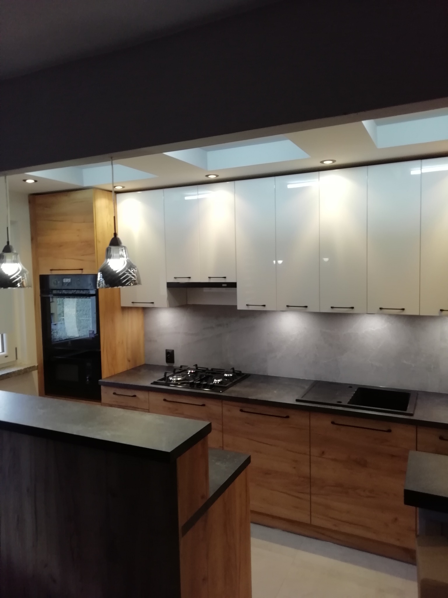 Nowoczesna kuchnia z drewnianymi szafkami dolnymi i białymi górnymi, czarnym blatem, oświetleniem podszafkowym i lampami wiszącymi w stylu industrialnym, widok na wyspę kuchenną.