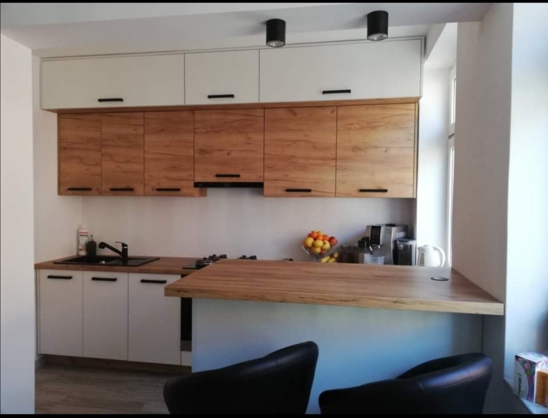 Jasna kuchnia z drewnianymi frontami szafek górnych i białymi dolnymi, czarne uchwyty i oświetlenie punktowe, drewniany blat z barkiem i dwa czarne hokery.