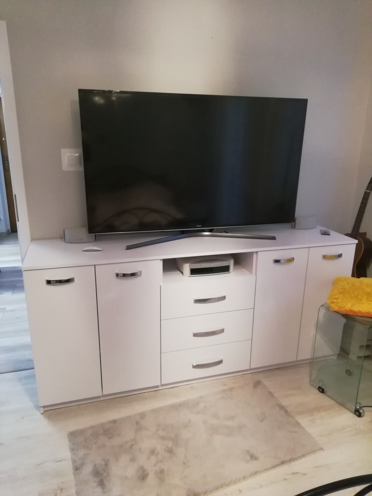 Biała szafka RTV z szufladami i szafkami, na której stoi telewizor; w tle widoczny fragment gitary.