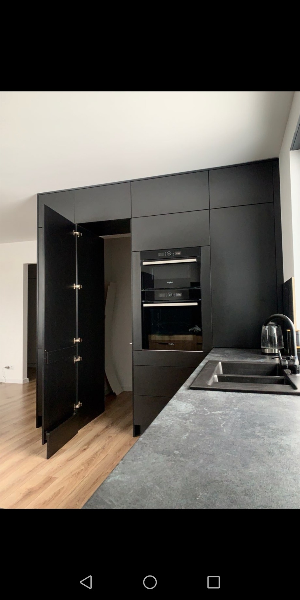 Ciemna zabudowa kuchenna z otwartymi drzwiczkami szafki, w której widać przestrzeń garderoby, obok piekarnik i czajnik elektryczny na blacie o teksturze kamienia.