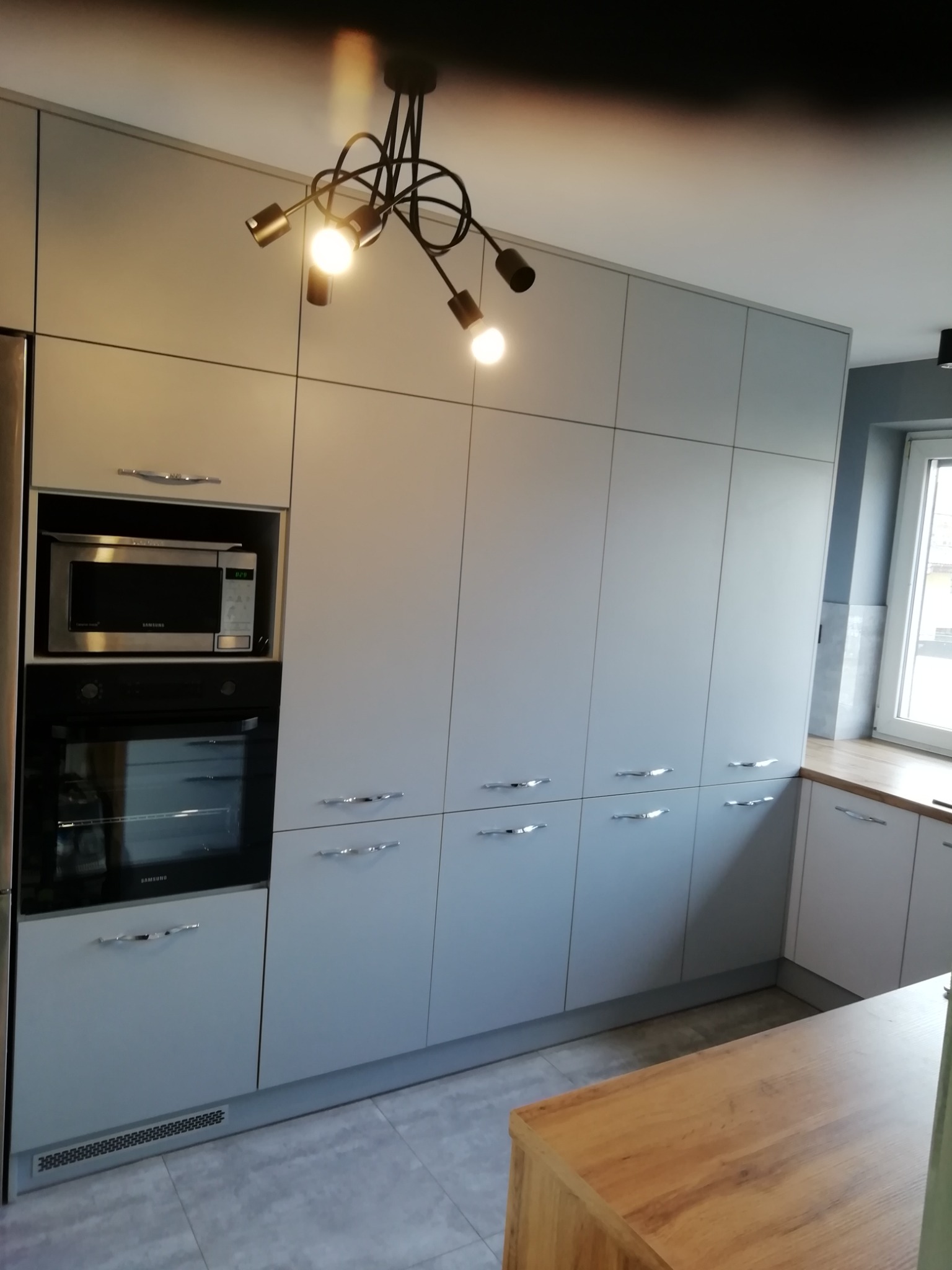 Jasnoszare szafki kuchenne z wbudowanym piekarnikiem i mikrofalówką, oświetlone nowoczesną lampą z kilkoma żarówkami