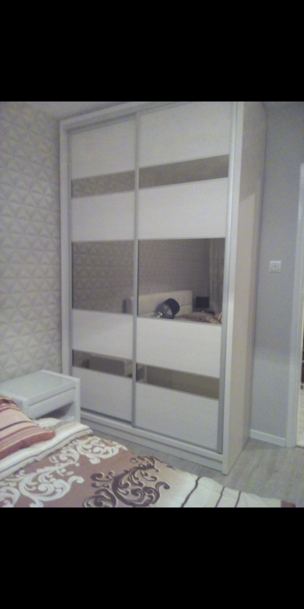 Biała szafa przesuwna z lustrami i dekoracyjnymi panelami w sypialni z wzorzystą tapetą i łóżkiem z brązową pościelą.