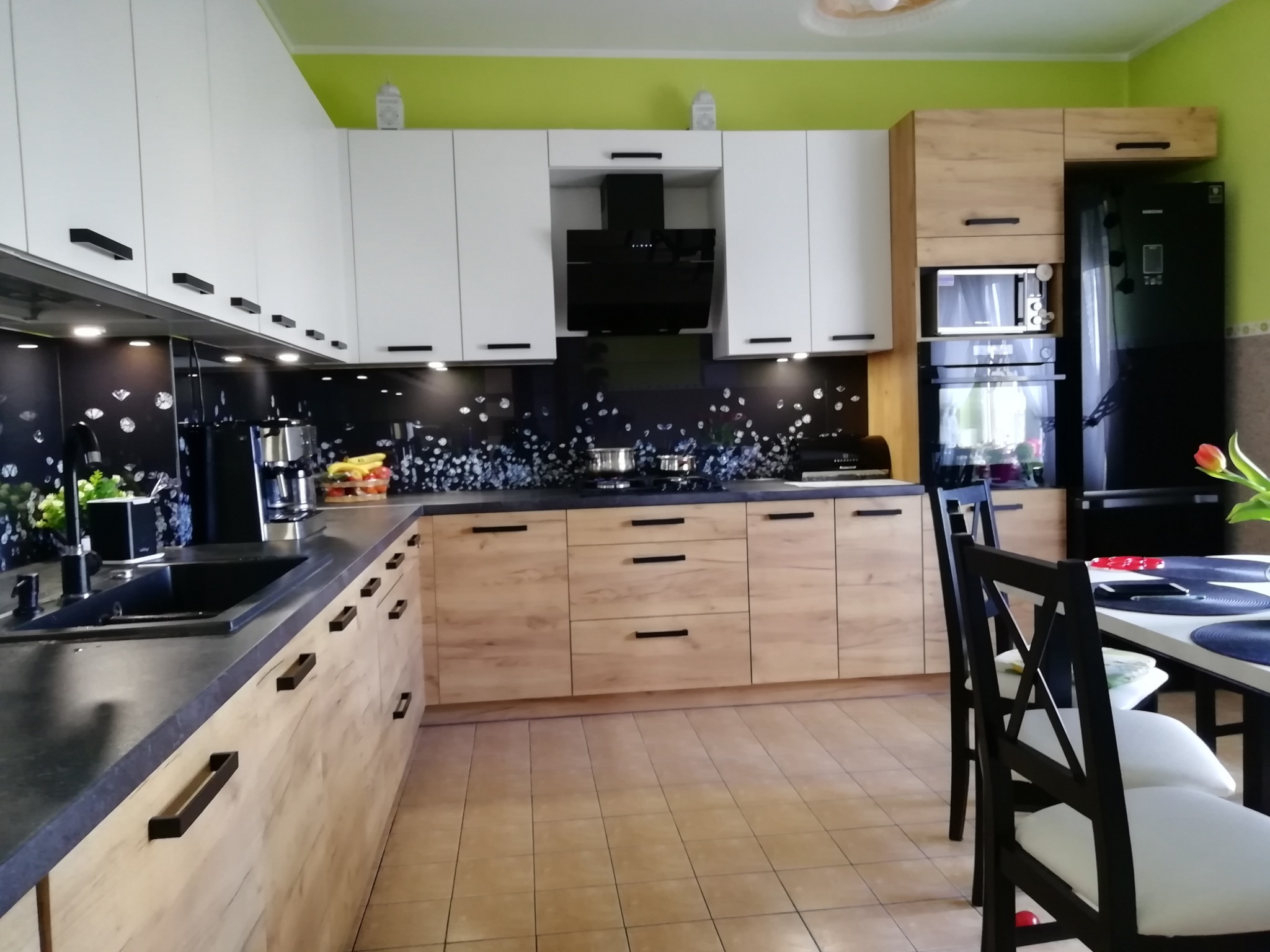 Nowoczesna kuchnia w Izbicach z frontami w kolorze jasnego drewna i białymi szafkami górnymi, czarny blat i sprzęt AGD, widoczne elementy dekoracyjne na blacie i stole.