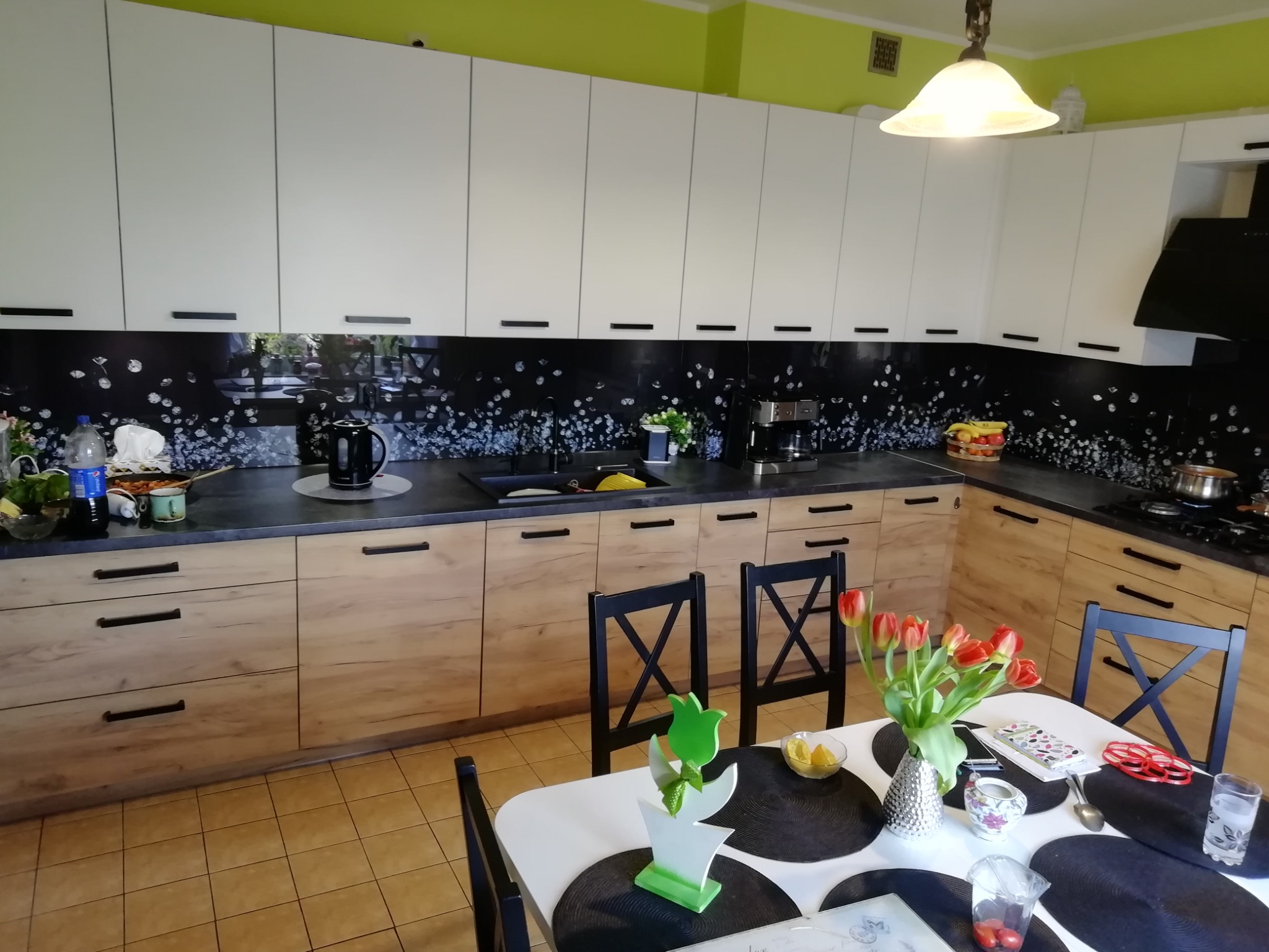 Jasna kuchnia z drewnianymi dolnymi szafkami i białymi górnymi, czarny panel szklany między szafkami, stół z tulipanami na pierwszym planie.