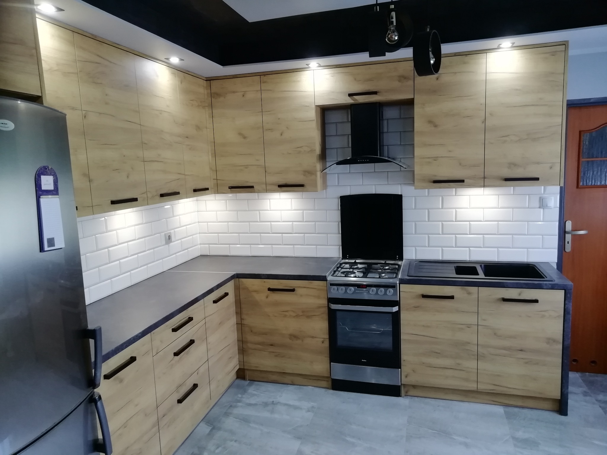 Jasna kuchnia z drewnopodobnymi frontami, ciemnymi blatami i białymi kafelkami na ścianie, wyposażona w srebrną lodówkę i kuchenkę gazową.