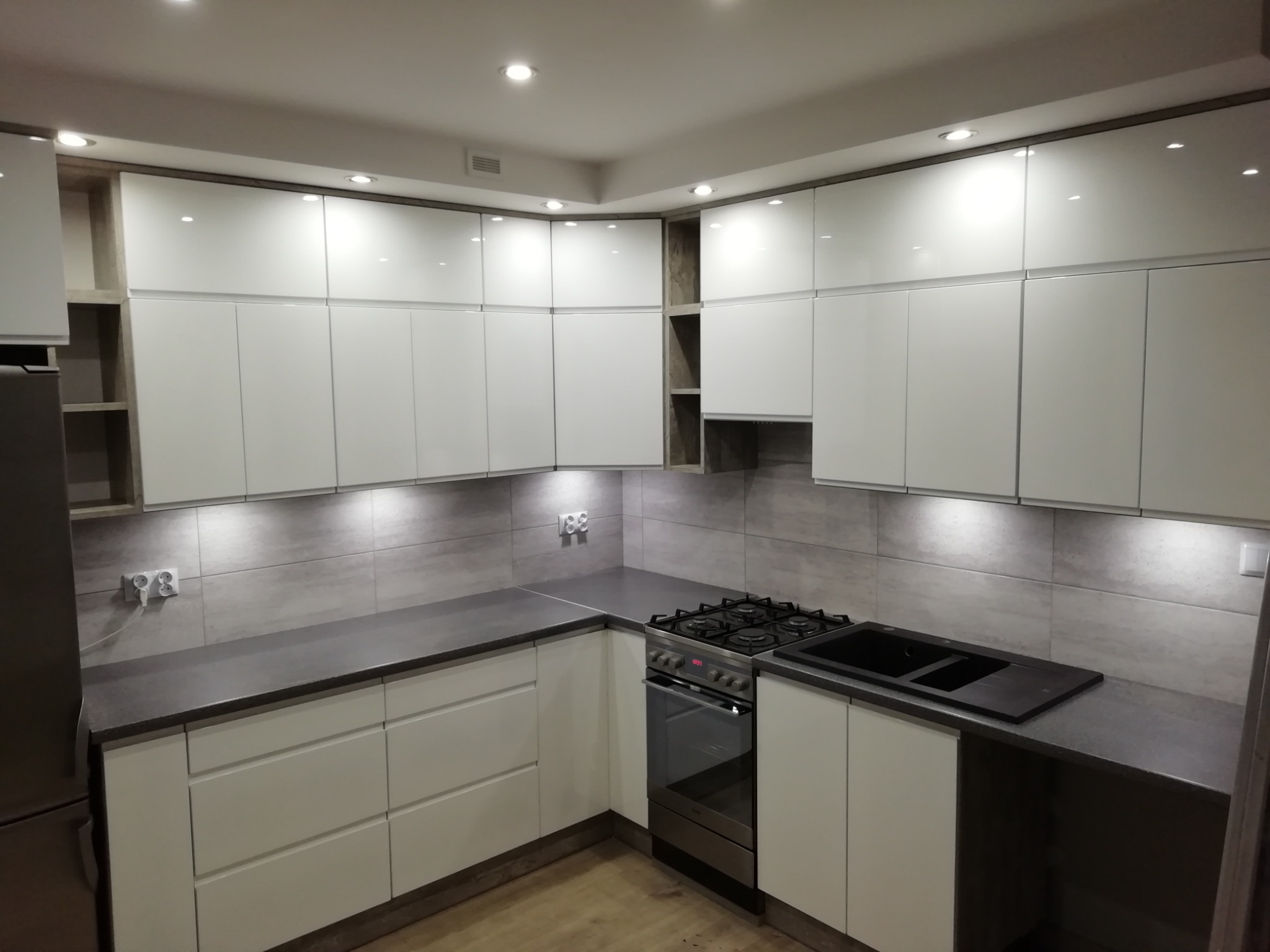 Nowoczesna, narożna kuchnia z białymi, lakierowanymi frontami, szarym blatem i płytkami imitującymi beton, oświetlona punktowymi lampami sufitowymi i listwą LED pod szafkami górnymi, z widoczną...