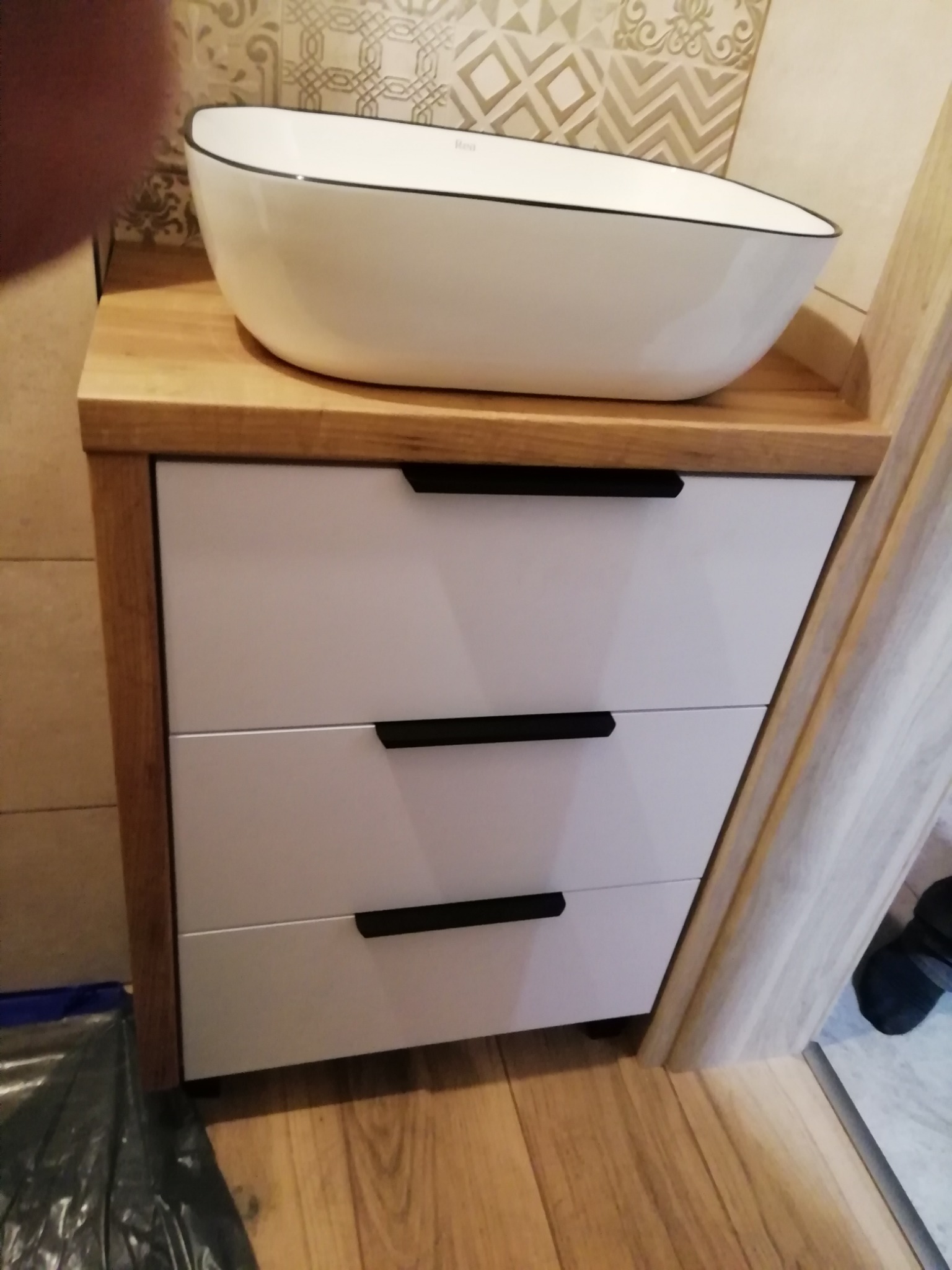 Biała umywalka nablatowa na szafce z trzema szufladami o białych frontach i drewnianym blacie, na tle wzorzystych płytek.