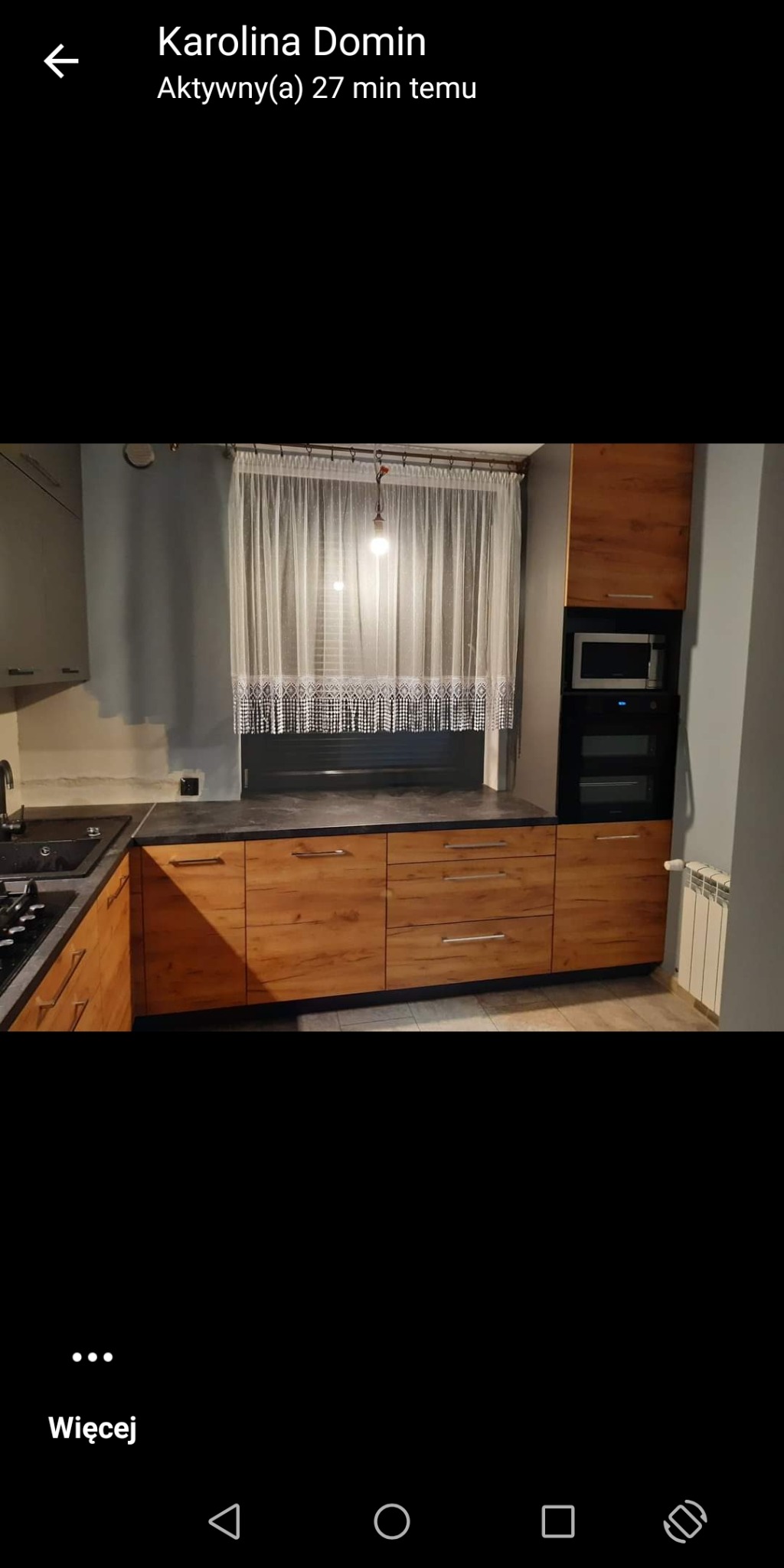 Nowoczesna kuchnia z drewnianymi frontami szafek, czarnym blatem i białymi firankami w oknie, widok na zabudowany piekarnik i mikrofalówkę.