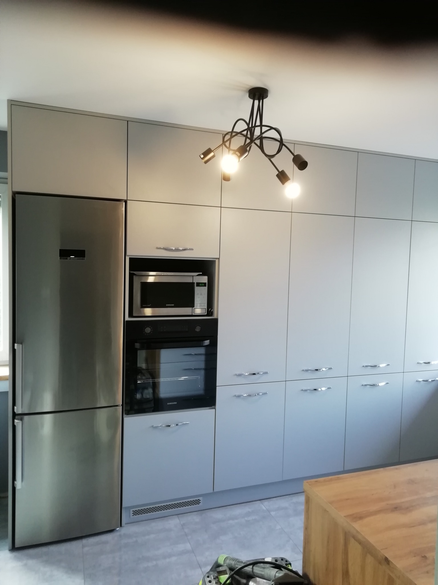 Nowoczesna kuchnia w odcieniach szarości z zabudową od podłogi do sufitu, srebrna lodówka side-by-side, piekarnik i mikrofalówka w zabudowie, oświetlenie sufitowe z czarnymi ramionami i drewniany...