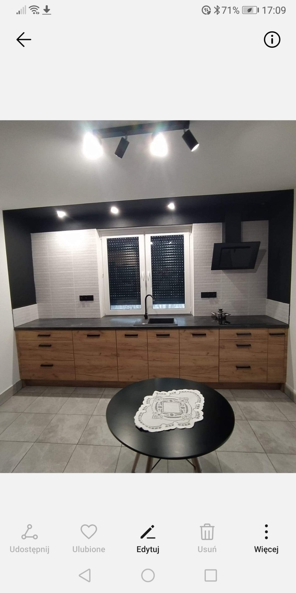 Nowoczesna kuchnia z drewnianymi frontami szafek, czarnym blatem i czarnym akcentem na ścianie, oświetlona reflektorami, z okrągłym stołem w tle.