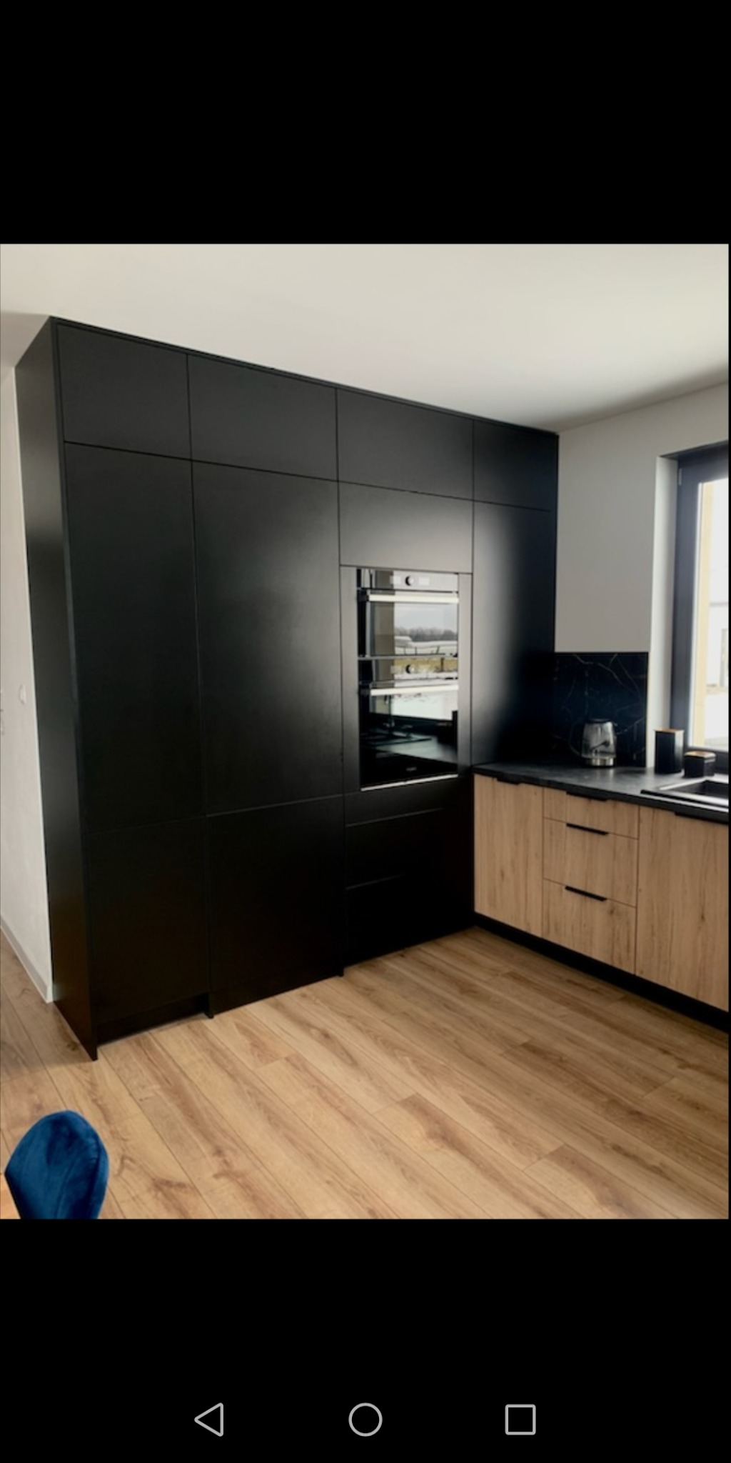 Nowoczesna kuchnia: wysoka zabudowa z czarnymi frontami, piekarnik w zabudowie, blaty drewnopodobne, czarny backsplash imitujący marmur, jasna drewniana podłoga.