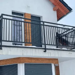 Czarna, metalowa balustrada balkonowa na tle białej elewacji budynku z drewnianymi akcentami w zimowej scenerii.