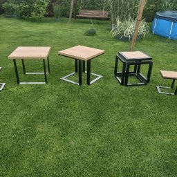 Emil Gradzik - Zestaw pięciu stolików i stołków z drewnianymi blatami i czarnymi metalowymi nogami o różnych kształtach, ustawionych na trawniku w ogrodzie z basenem w tle.