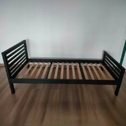 Emil Gradzik - Czarna metalowa rama łóżka z drewnianym stelażem listwowym, ustawiona na podłodze z paneli, na tle białej ściany.