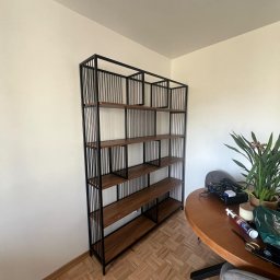 Emil Gradzik - Metalowy regał z drewnianymi półkami, czarna rama z pionowymi prętami, stoi przy białej ścianie, obok okrągły drewniany stół z rośliną doniczkową i różnymi przedmiotami.