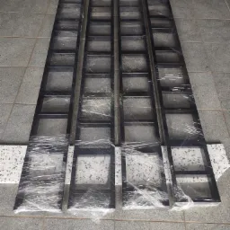 Cztery elementy metalowej konstrukcji, prawdopodobnie schodów lub balustrady, zabezpieczone folią stretch na szarym, płytkowym podłożu. Widoczne fragmenty białych podkładek pod elementami.