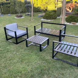 Zestaw mebli ogrodowych: fotel z szarą poduszką, ławka i stolik kawowy z metalową konstrukcją i drewnianymi szczebelkami, ustawione na trawniku, w tle krzewy i drzewa.