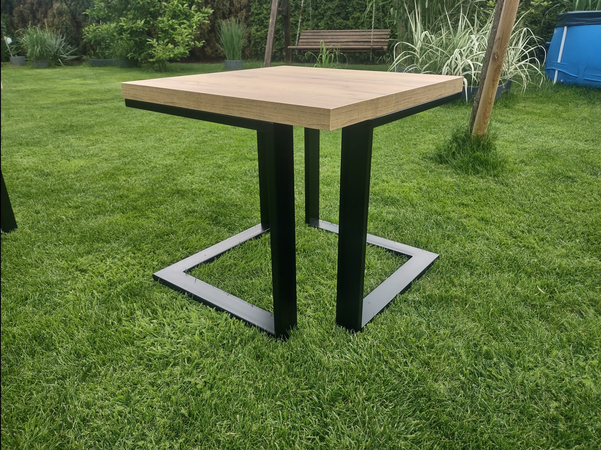 Stół ogrodowy z drewnianym blatem i czarną, metalową podstawą w kształcie kwadratu, ustawiony na trawniku w Lubinie. W tle widoczny basen i huśtawka.