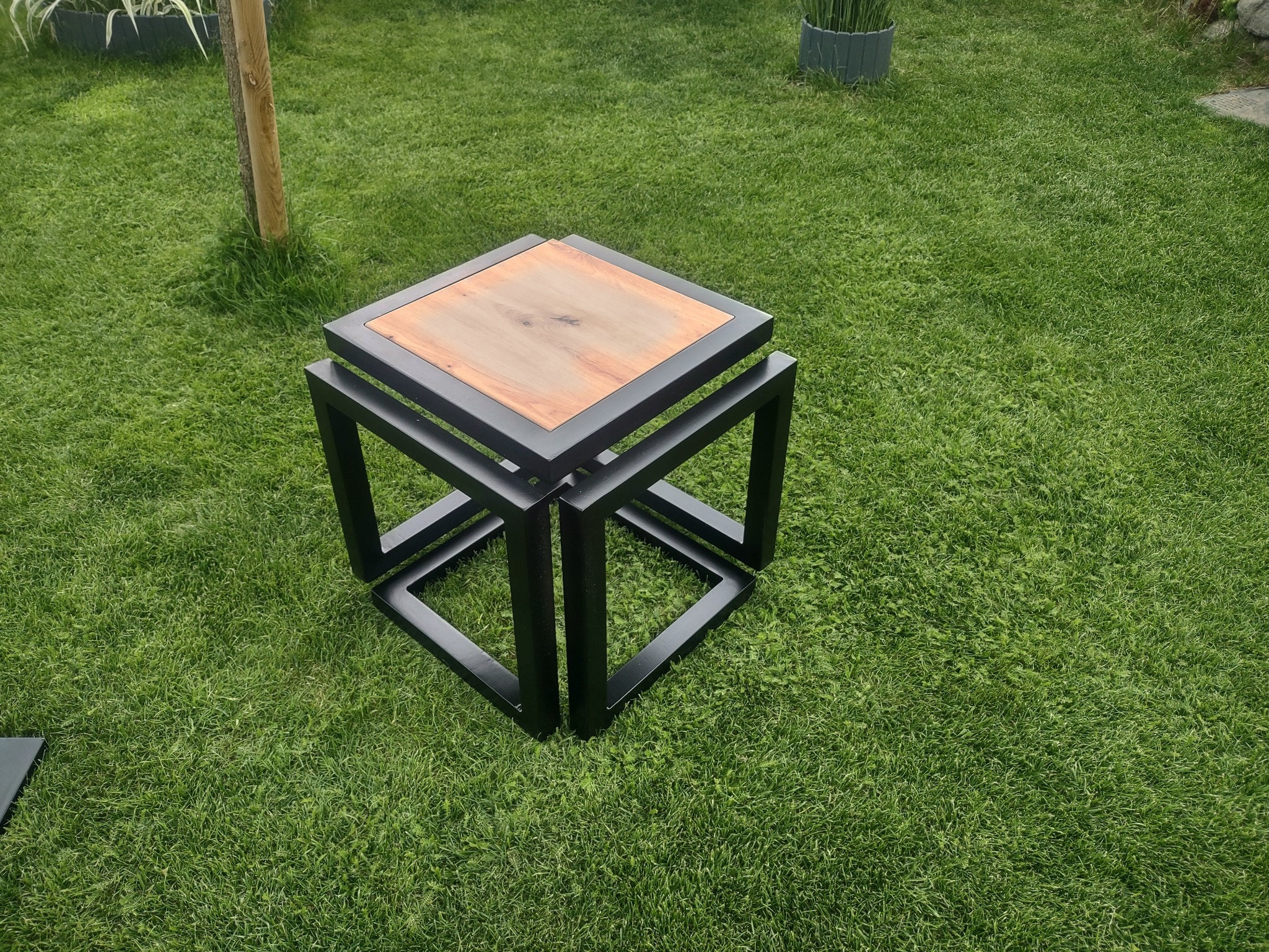 Kwadratowy stolik kawowy z drewnianym blatem i czarną, metalową ramą, umieszczony na trawniku.