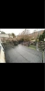 Czarna, pozioma konstrukcja ogrodzenia z metalowych profili na tle asfaltowej powierzchni i zabudowań. Widok z góry, lekko z boku.