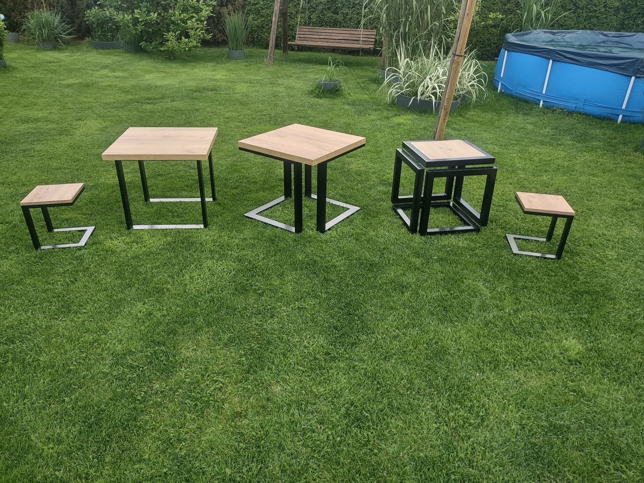 Zestaw pięciu stolików i stołków z drewnianymi blatami i czarnymi metalowymi nogami o różnych kształtach, ustawionych na trawniku w ogrodzie z basenem w tle.