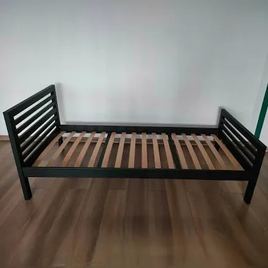 Czarna metalowa rama łóżka z drewnianym stelażem listwowym, ustawiona na podłodze z paneli, na tle białej ściany.