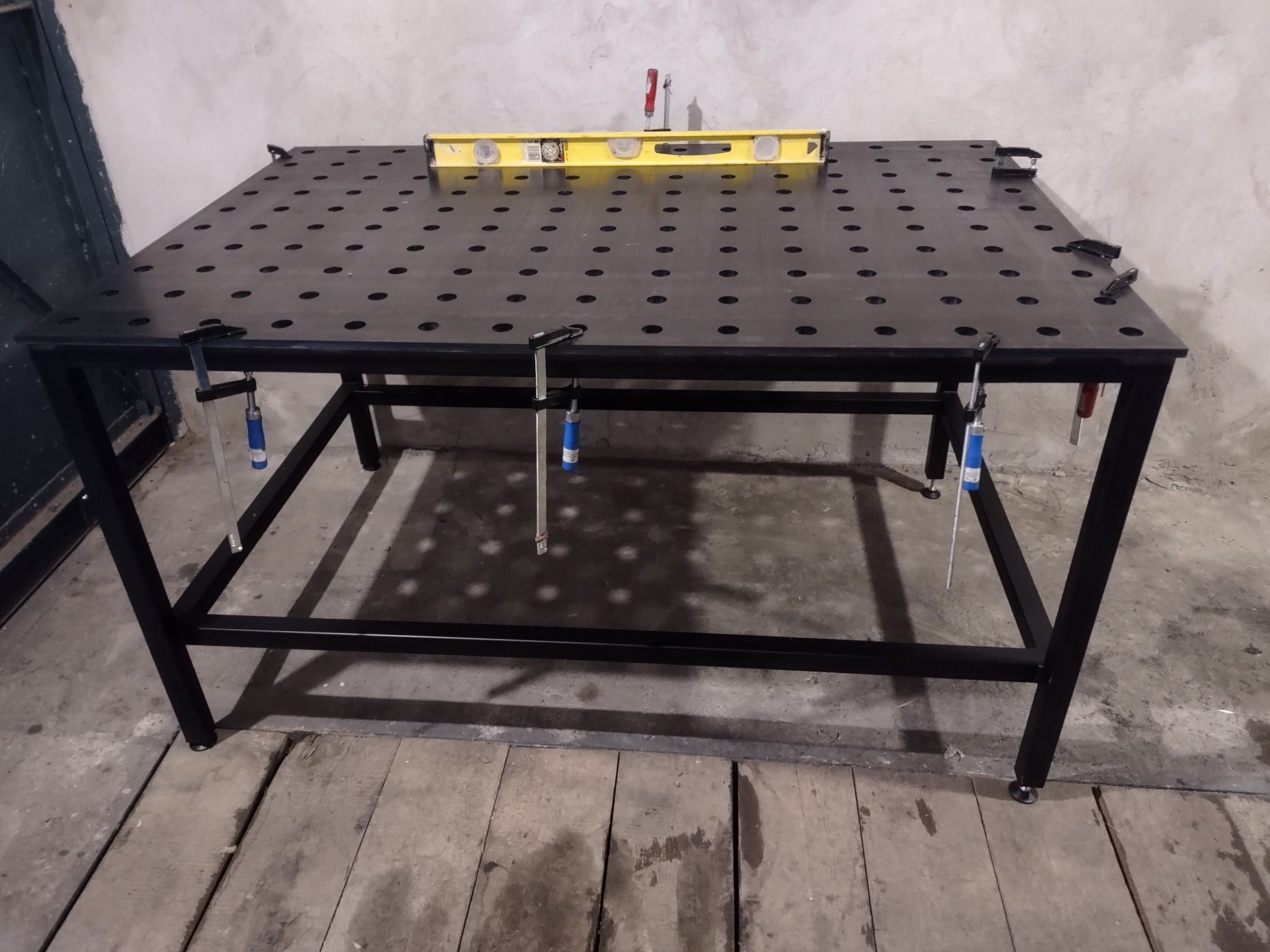 Stół spawalniczy z perforowanym blatem, poziomica na blacie, widoczne zaciski śrubowe, ustawiony w warsztacie z betonową ścianą i drewnianą podłogą.