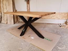 Stół z blatem z jasnego drewna i czarną, metalową podstawą w kształcie litery X, umieszczony na kartonie w warsztacie.