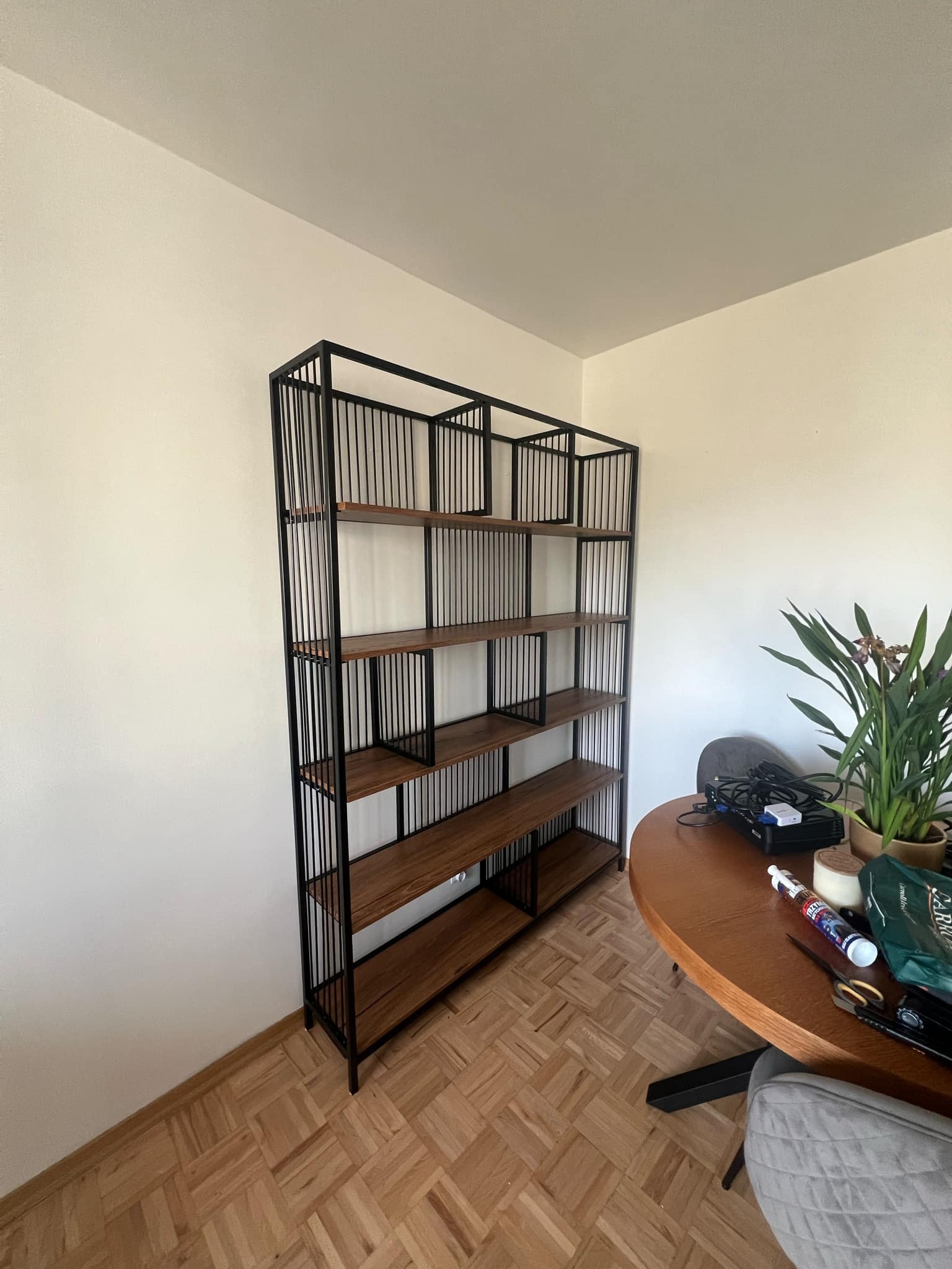 Metalowy regał z drewnianymi półkami, czarna rama z pionowymi prętami, stoi przy białej ścianie, obok okrągły drewniany stół z rośliną doniczkową i różnymi przedmiotami.