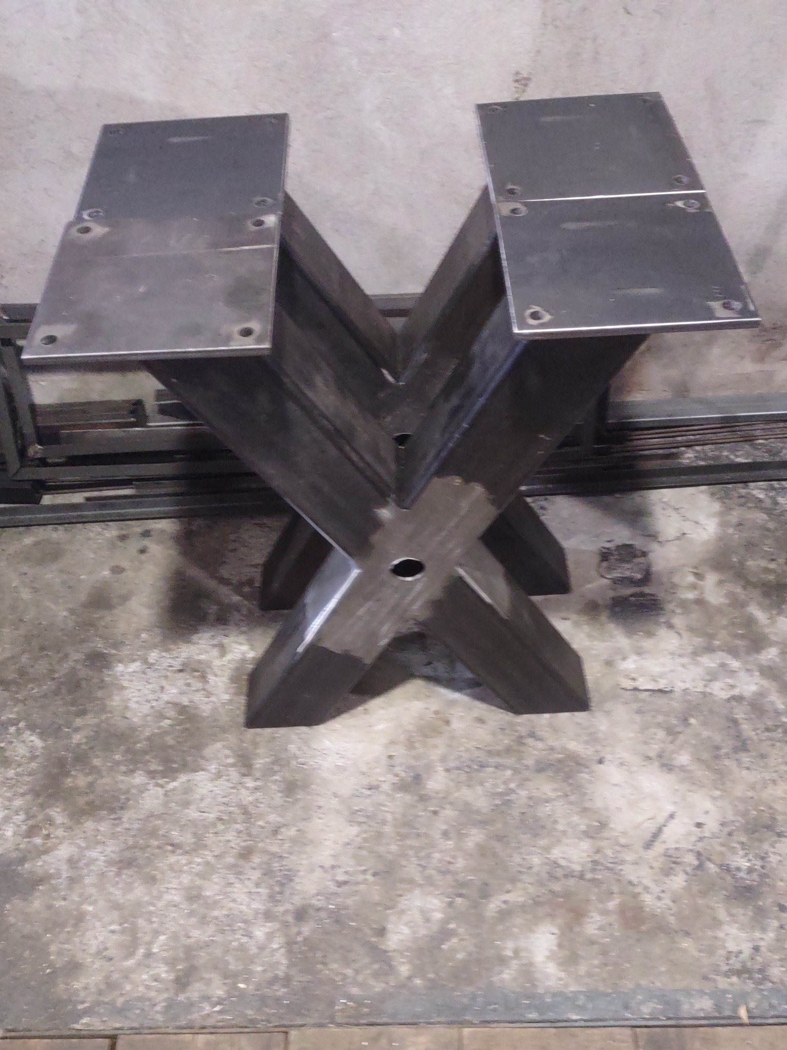 Surowa, spawana podstawa stołu z metalu w kształcie litery X, z przyspawanymi płytami montażowymi z otworami na śruby, widoczna na tle betonowej podłogi i elementów metalowych.