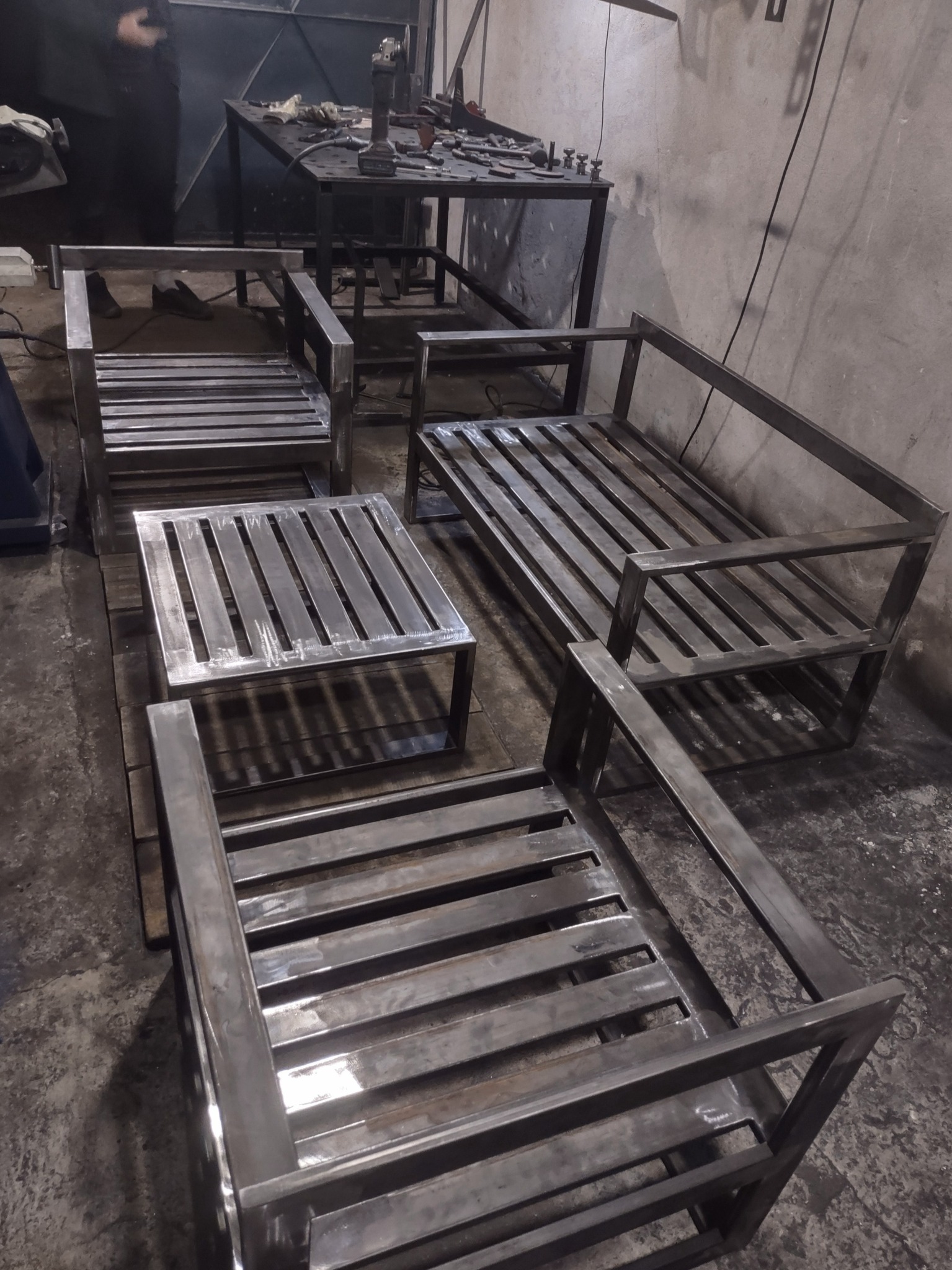 Surowe, metalowe ramy krzeseł i stolika kawowego w warsztacie, z widocznymi śladami spawania i obróbki, ustawione na podłodze w pobliżu stołu warsztatowego z narzędziami.