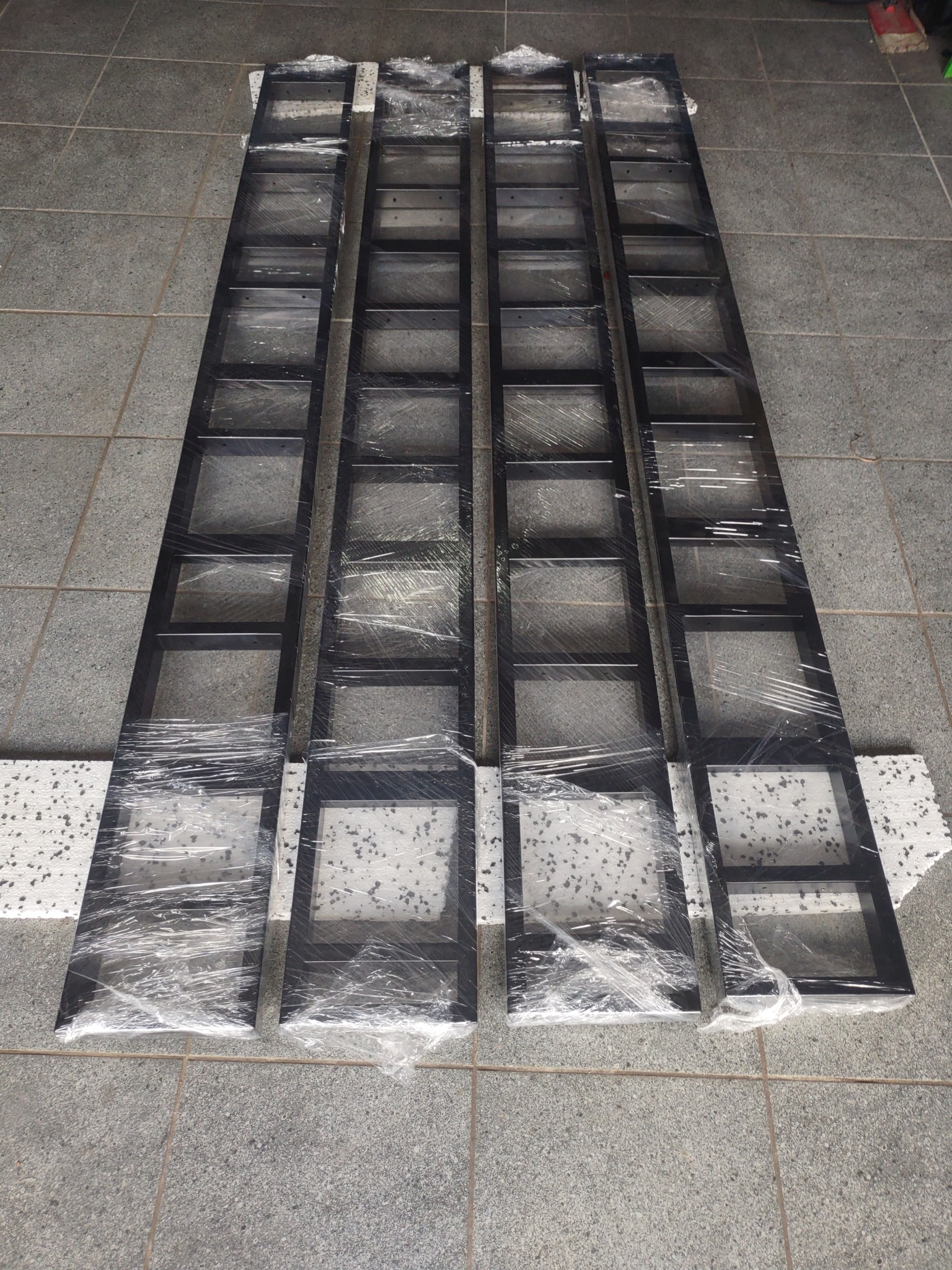 Cztery elementy metalowej konstrukcji, prawdopodobnie schodów lub balustrady, zabezpieczone folią stretch na szarym, płytkowym podłożu. Widoczne fragmenty białych podkładek pod elementami.