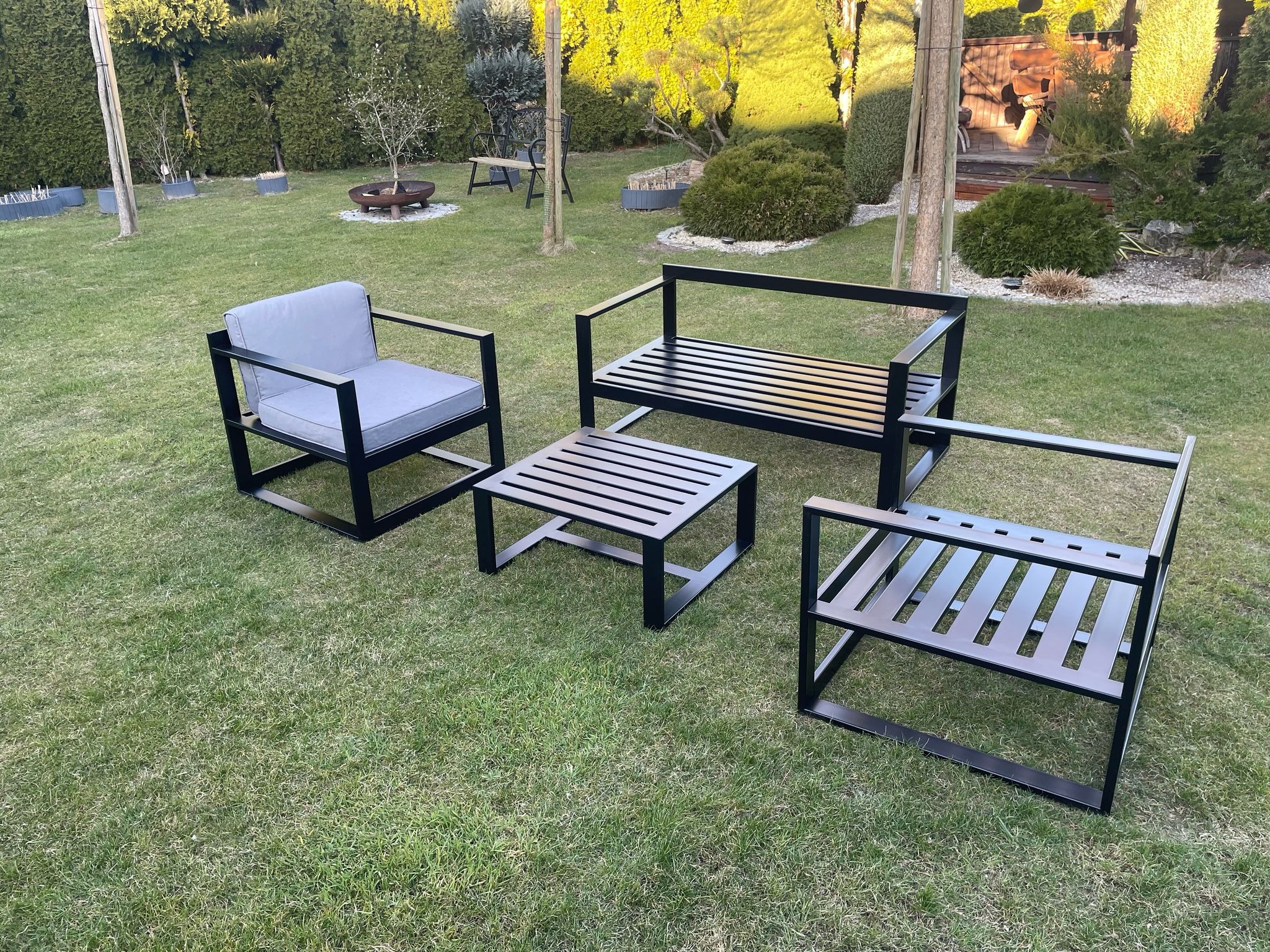Zestaw mebli ogrodowych: fotel z szarą poduszką, ławka i stolik kawowy z metalową konstrukcją i drewnianymi szczebelkami, ustawione na trawniku, w tle krzewy i drzewa.
