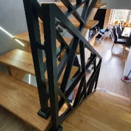 Drewniane schody z czarną, geometryczną balustradą, podświetlone ledami na szarej ścianie, widok z góry na dół.