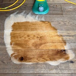 Artur Trembiński - Fragment podłogi z desek, częściowo odnowionej, z widoczną maszyną do cyklinowania i kablem zasilającym. Kontrast między starą a odnowioną powierzchnią.