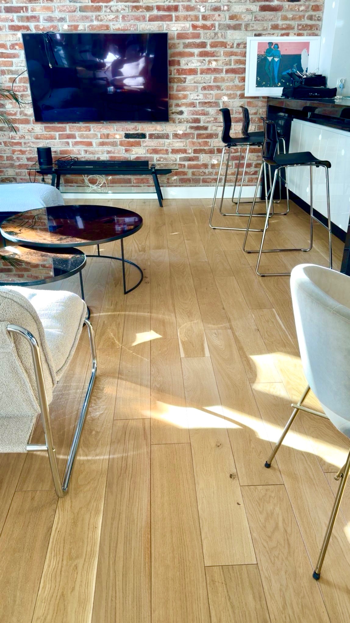 Jasne, drewniane panele podłogowe w salonie z ceglaną ścianą i nowoczesnymi meblami. Widoczne subtelne refleksy światła na podłodze.