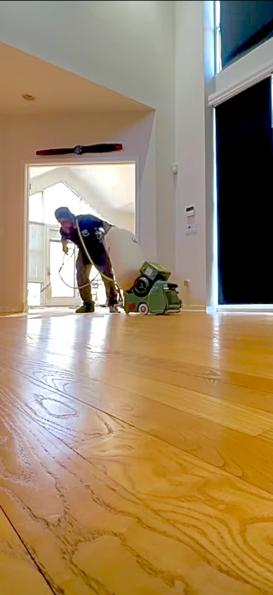 Pracownik w trakcie cyklinowania parkietu w jasnym wnętrzu. Widoczna maszyna do cyklinowania i worek na pył. Na ścianie dekoracja - śmigło samolotu.