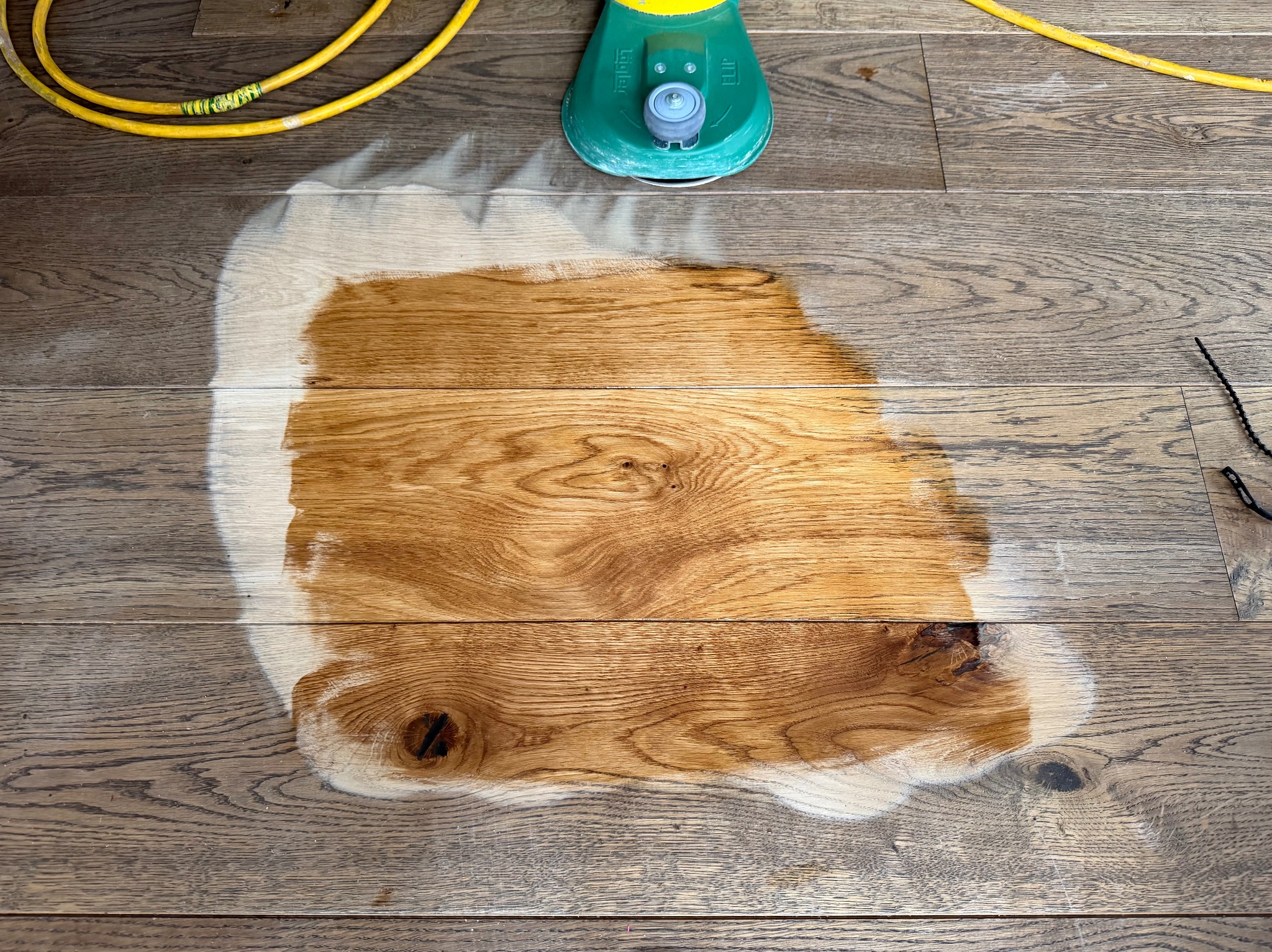 Fragment podłogi z desek, częściowo odnowionej, z widoczną maszyną do cyklinowania i kablem zasilającym. Kontrast między starą a odnowioną powierzchnią.