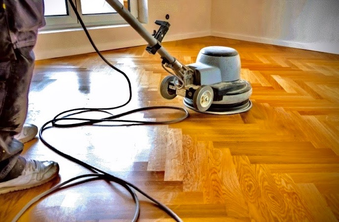 Maszyna do renowacji parkietu w trakcie pracy w Warszawie. Widoczny fragment podłogi w jodełkę po obróbce, z wyraźnym połyskiem. Fragment osoby obsługującej maszynę.