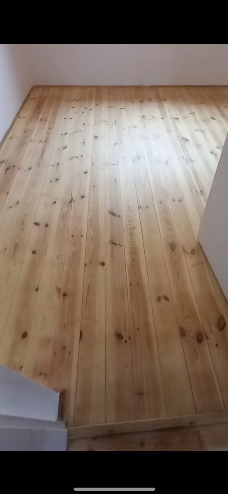 Jasne, odnowione deski podłogowe z widocznymi słojami drewna, ułożone wzdłuż pomieszczenia. Świeżo wykończona podłoga w pokoju z białymi ścianami.