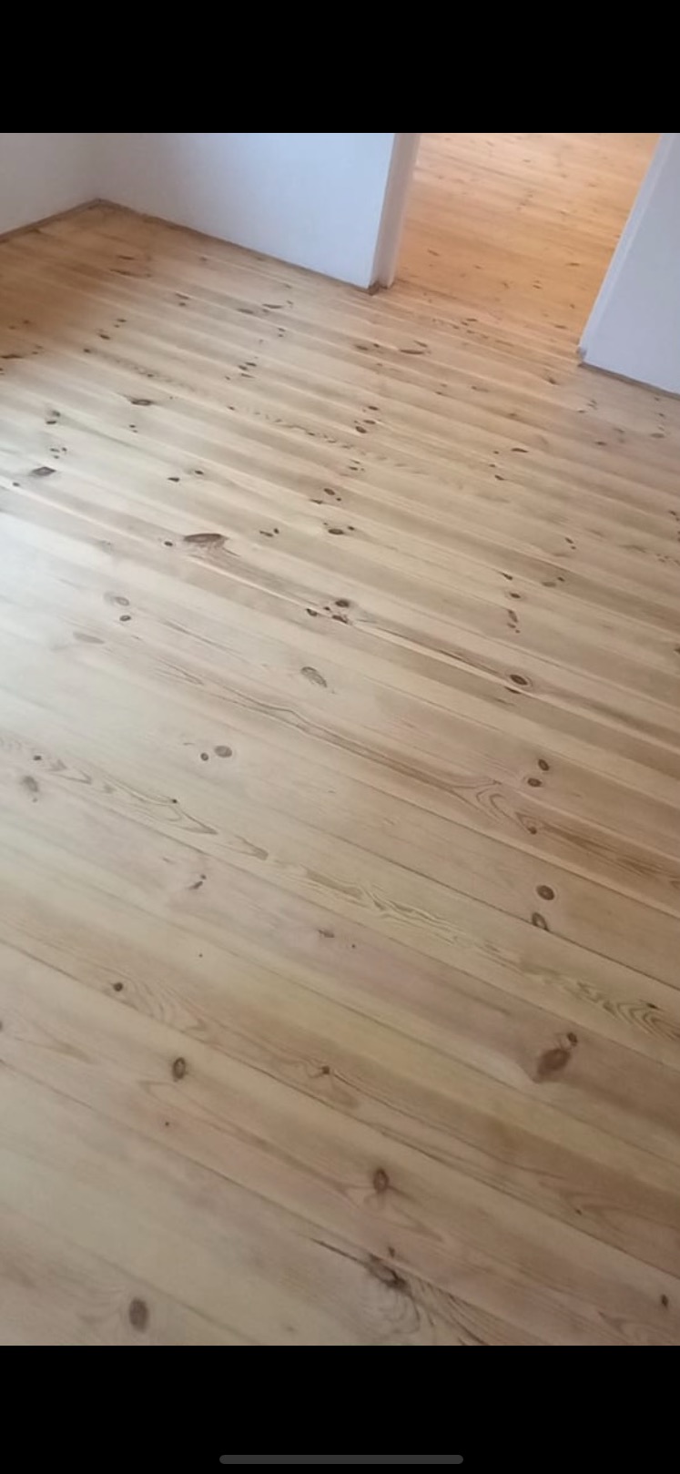 Jasna, odnowiona podłoga z desek z widocznymi słojami i sękami, prowadząca do otwartego przejścia między pomieszczeniami. Naturalne światło podkreśla strukturę drewna.