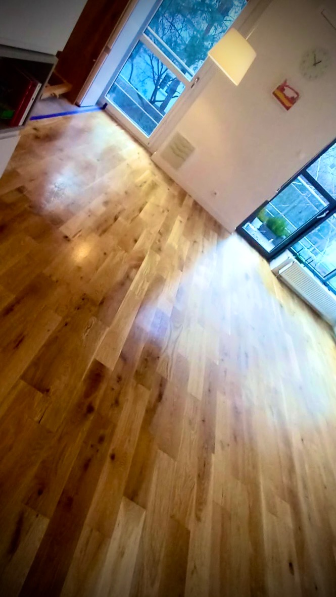 Jasny parkiet ułożony w jodełkę francuską, widok z góry, z oknami w tle. Naturalne oświetlenie podkreśla strukturę drewna i dodaje ciepła wnętrzu.