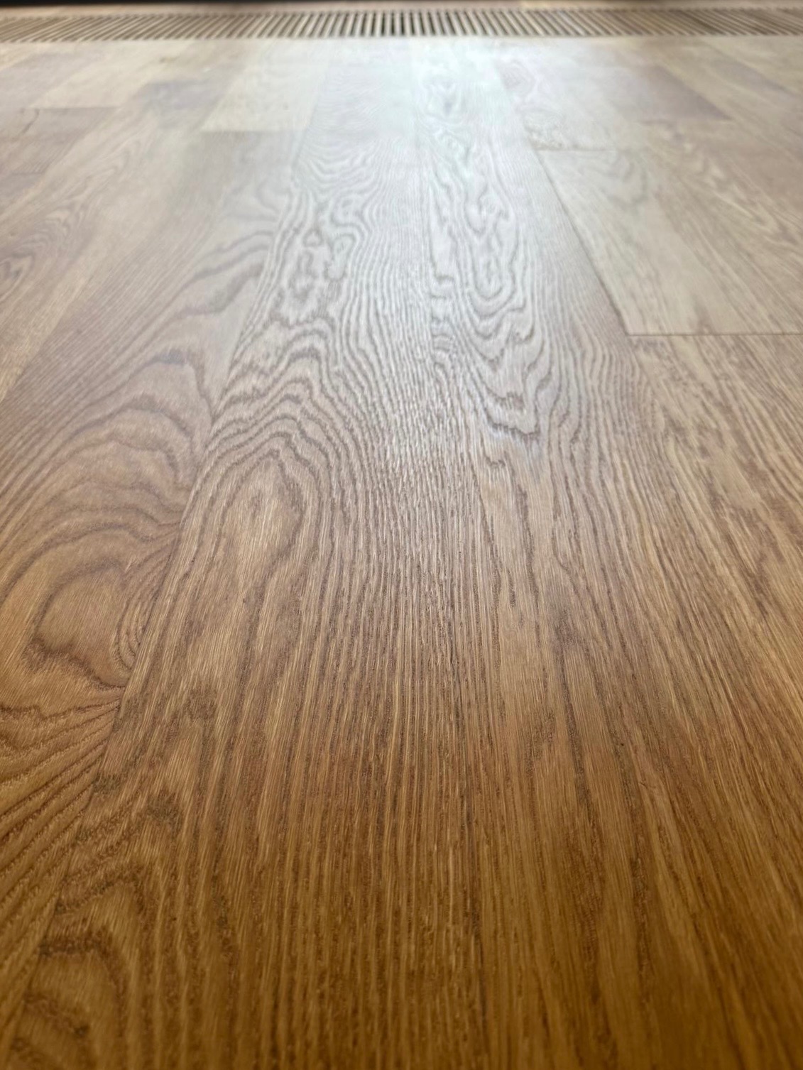 Ułożone panele podłogowe w odcieniach brązu, z widocznym wzorem drewna i grzejnikiem w tle. Perspektywa z niskiego kąta podkreśla strukturę podłogi.