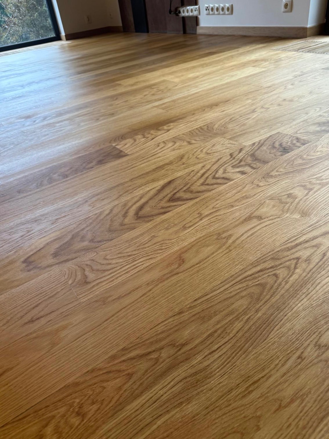 Błyszcząca, świeżo wykończona podłoga z parkietu dębowego ułożonego w jodełkę, z widocznymi słojami drewna i naturalnym oświetleniem wpadającym przez okno.