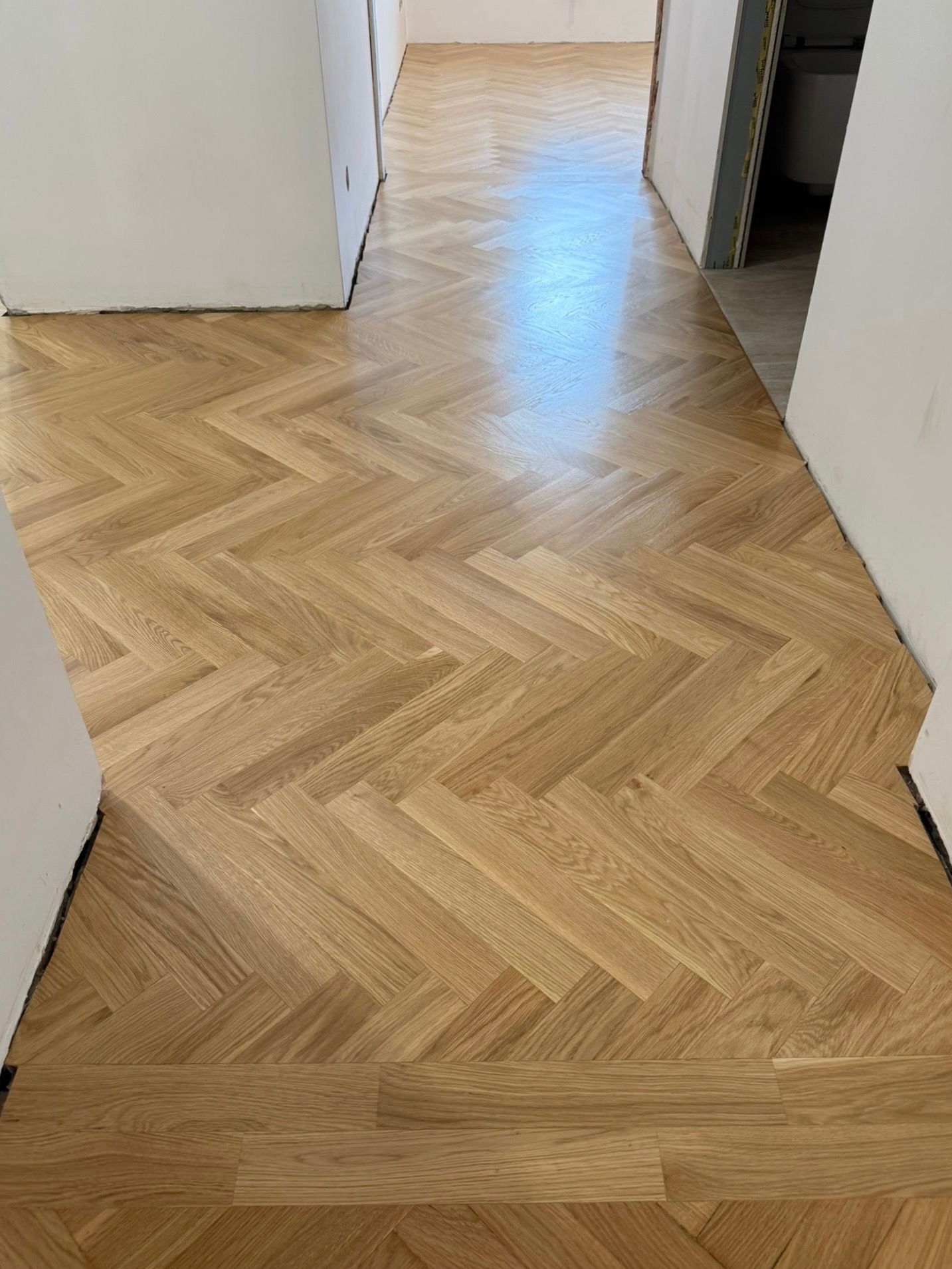 Ułożony parkiet w jodełkę, jasne drewno, widok z góry, fragment korytarza z białymi ścianami i otwartymi drzwiami w tle. Nowa podłoga.