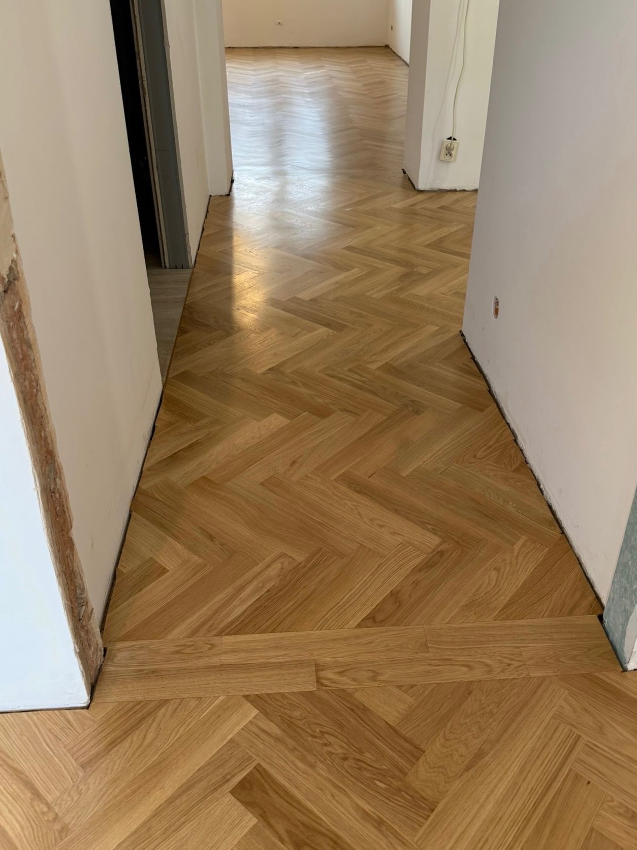 Jasny parkiet ułożony we wzór jodełki w korytarzu, widok z góry. Świeżo położona podłoga z drewna, widoczne przejście do kolejnych pomieszczeń. Białe ściany.
