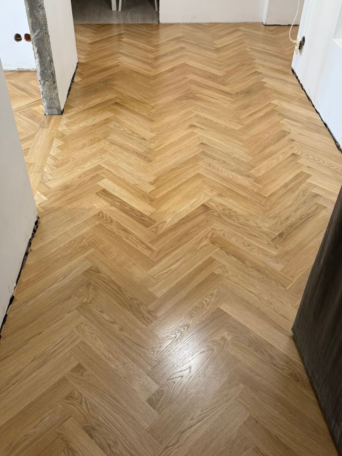 Jasny, parkiet ułożony w jodełkę, świeżo po renowacji. Widoczne detale drewna i wzór ułożenia. W tle fragmenty ścian i drzwi.