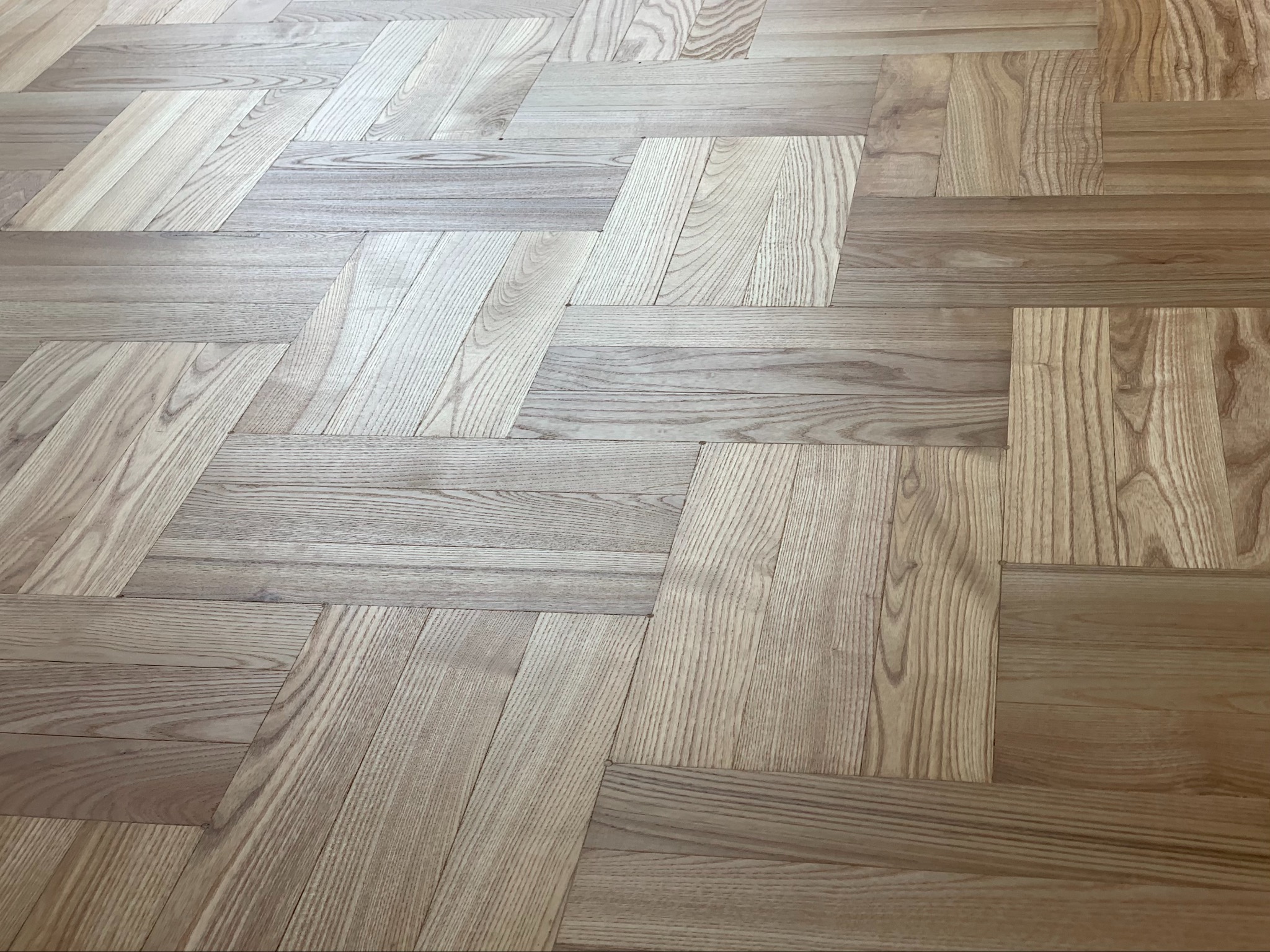 Jasna podłoga ułożona w jodełkę z parkietu o wyraźnym usłojeniu, widoczne różnice w odcieniach poszczególnych klepek.