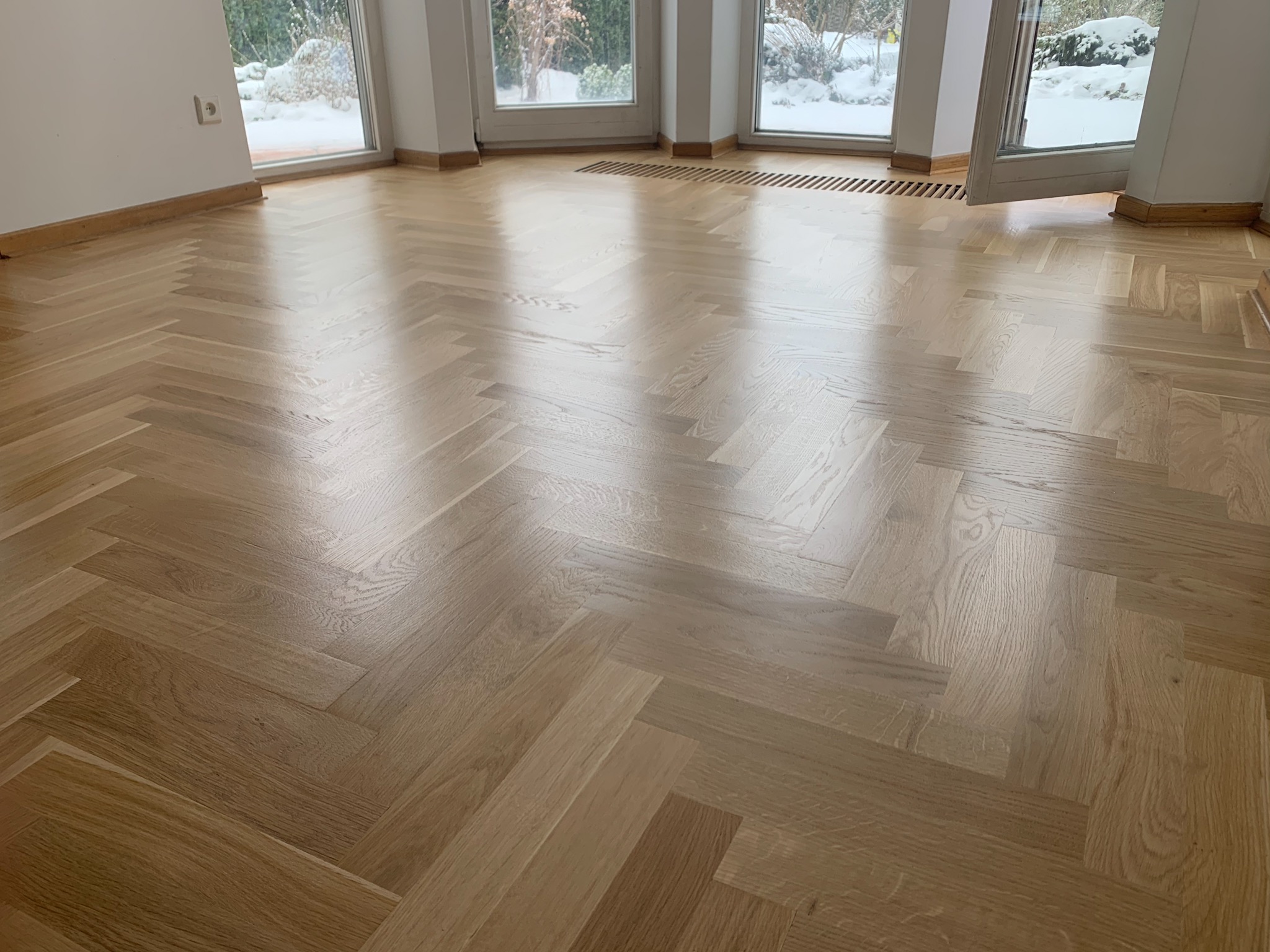 Jasna, lakierowana podłoga ułożona w jodełkę francuską w przestronnym pokoju z oknami wychodzącymi na zimowy ogród.