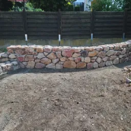 Niski murek oporowy z nieregularnych kamieni w odcieniach czerwieni i brązu, częściowo ukończony, z widocznymi śladami prac ziemnych i stertą kamieni obok, na tle drewnianego płotu.